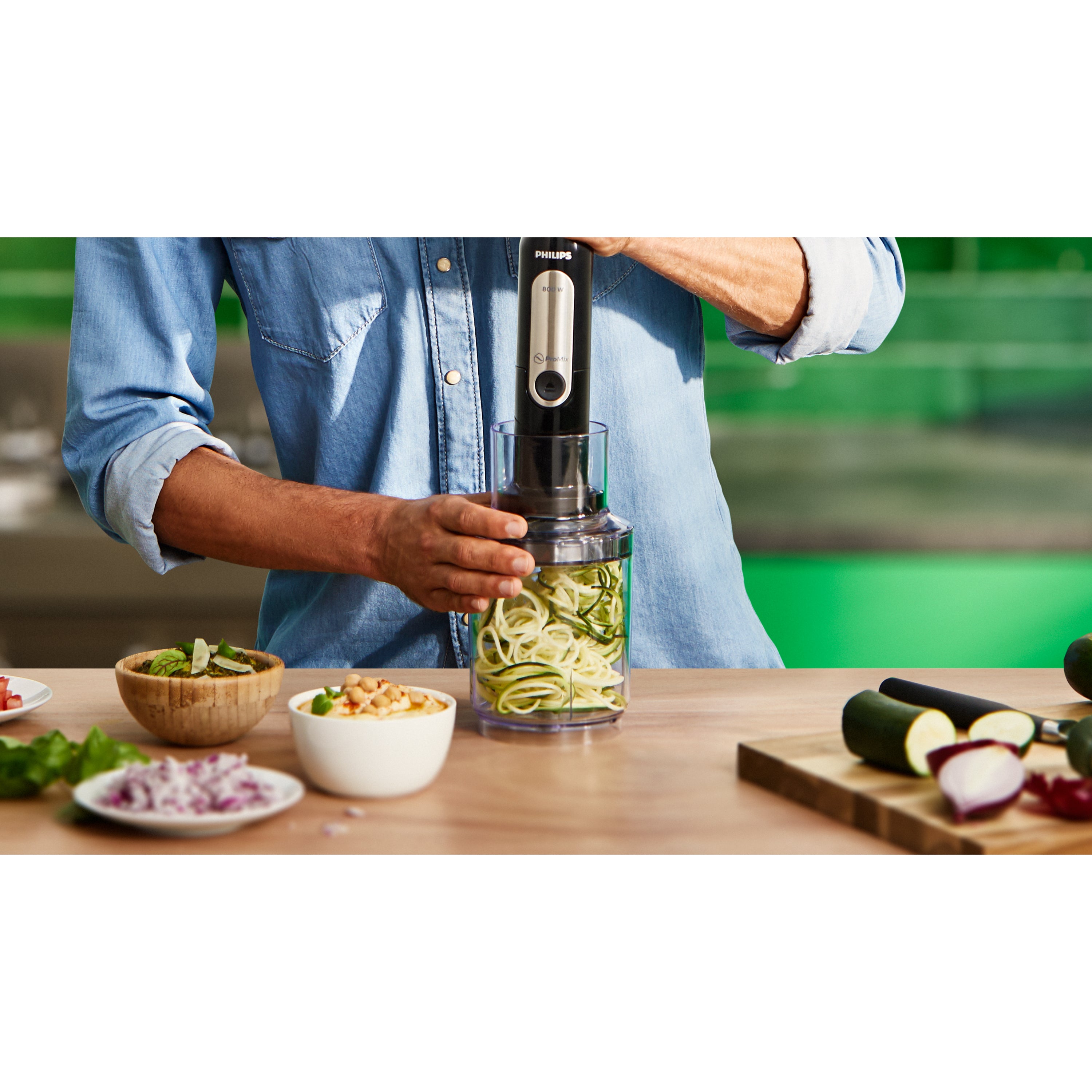 Person using a spiralizer to make zucchini noodles on a kitchen counter with various vegetables.