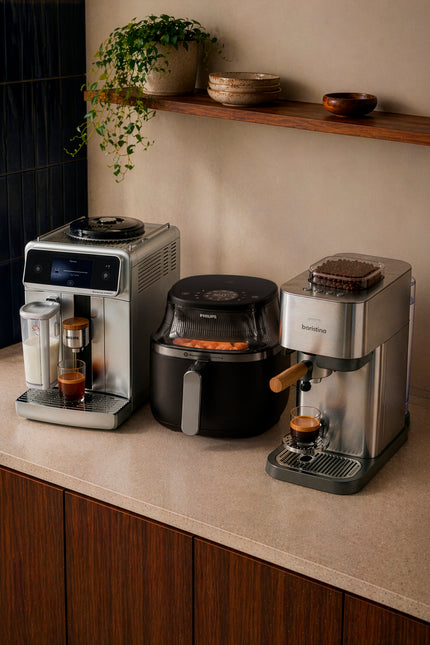 A countertop kitchen scene showing three appliances: a stainless steel coffee machine on the left, a black air fryer in the center, and a stainless steel espresso machine on the right, with a shelf and plants above.